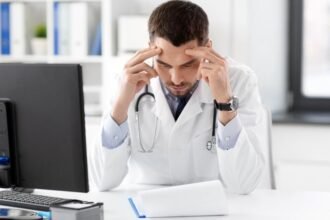 Stresszes orvos dolgozik az irodájában számítógép előtt.