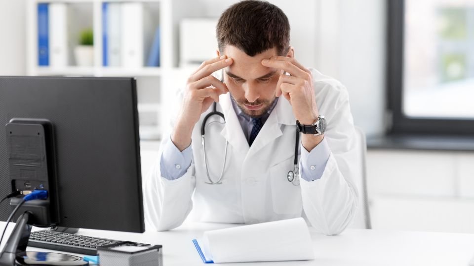 Stresszes orvos dolgozik az irodájában számítógép előtt.